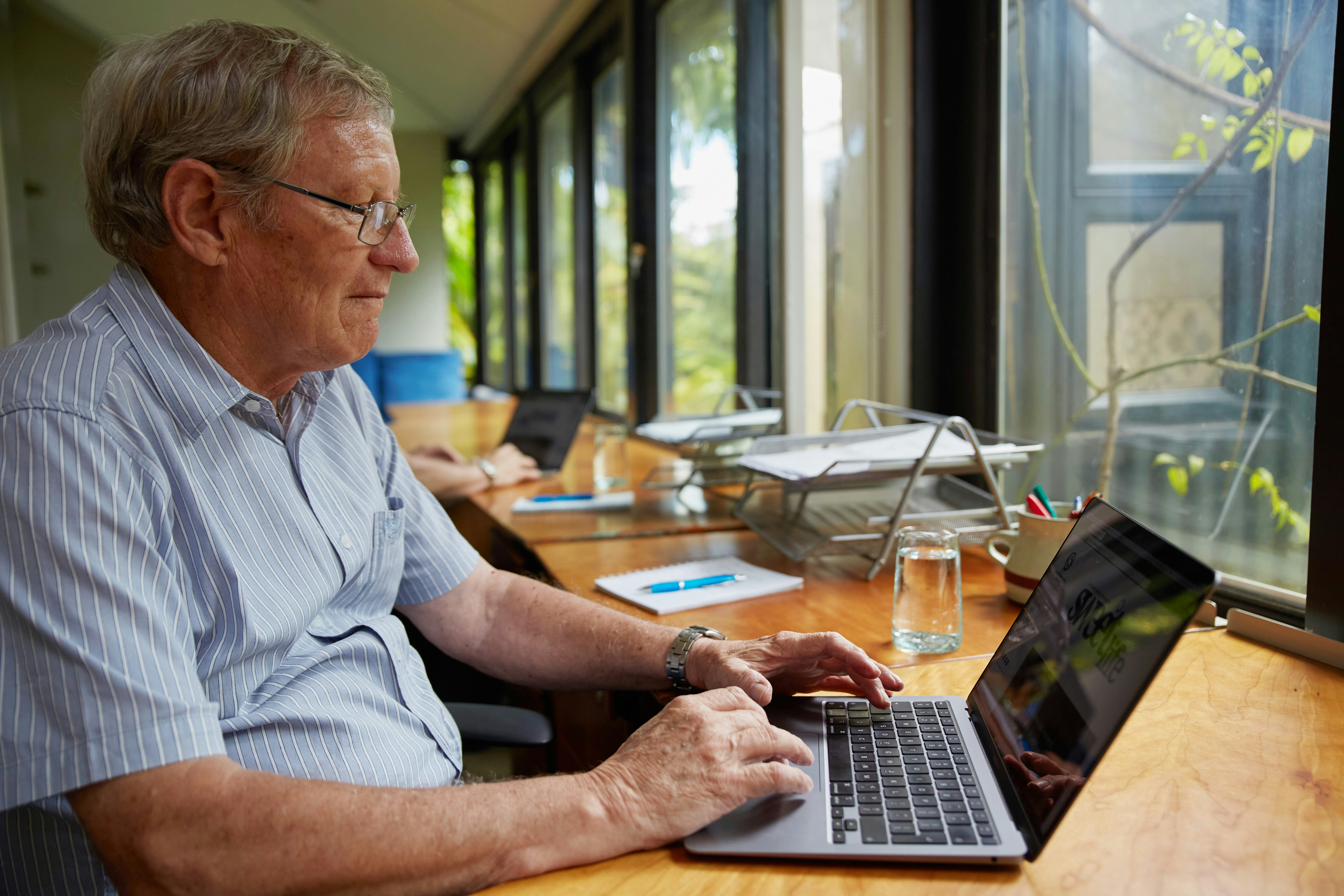 Senior citizen gentleman working on a laptop computer in a beautiful modern office with lots of natural light and foliage visible through the large windows. Photo by Sweet Life on Unsplash