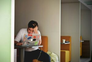 A man working on a laptop, holding his head in a gesture indicating anxiety and stress.