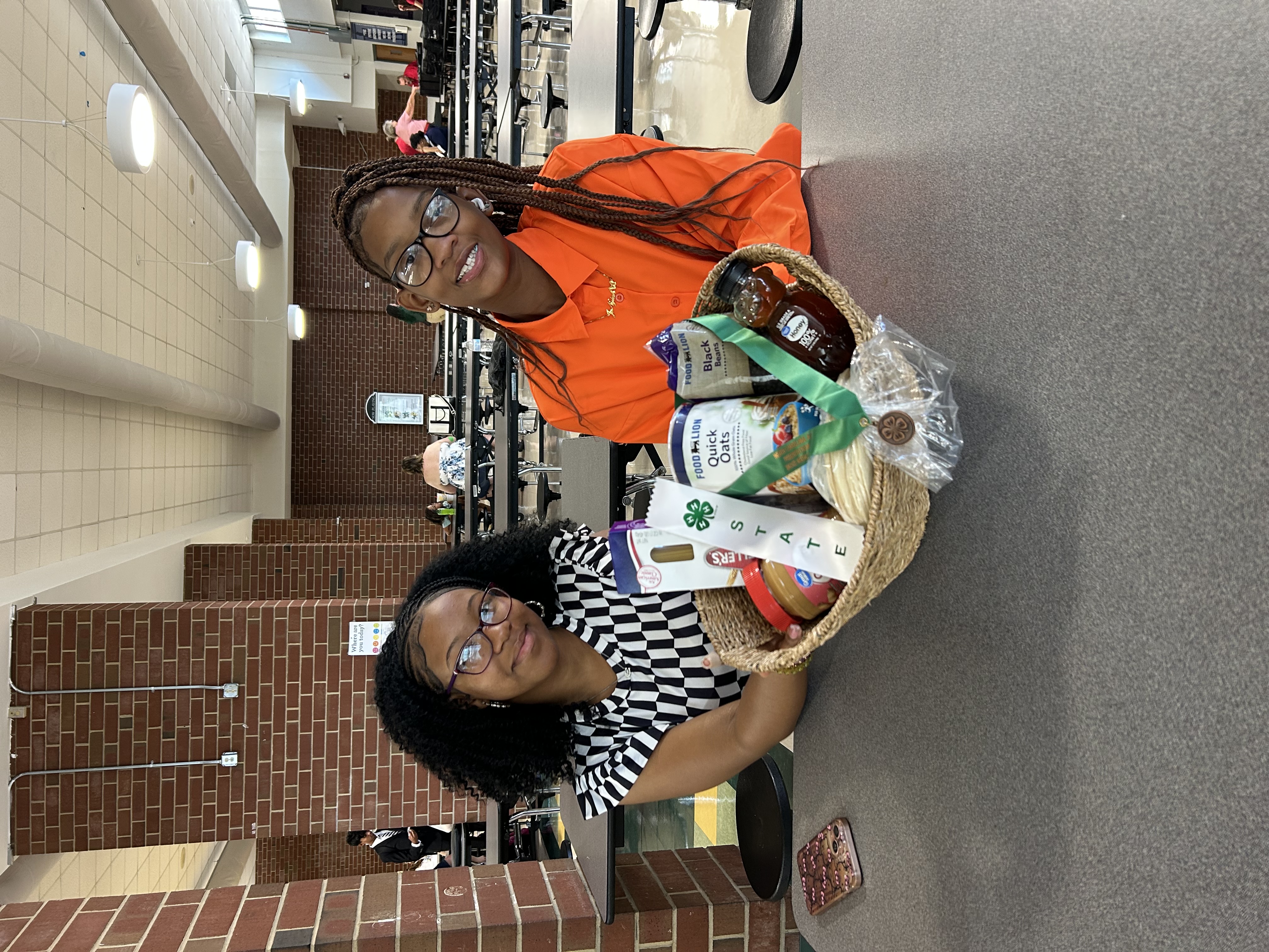 Two girls - one wearing an orange shirt and one wearing a black and white striped dress - sitting at a table in a cafeteria. A basket sits in front of them, filled with non-perishables, with a bronze medal and a white ribbon saying "State" in green letters resting on top.