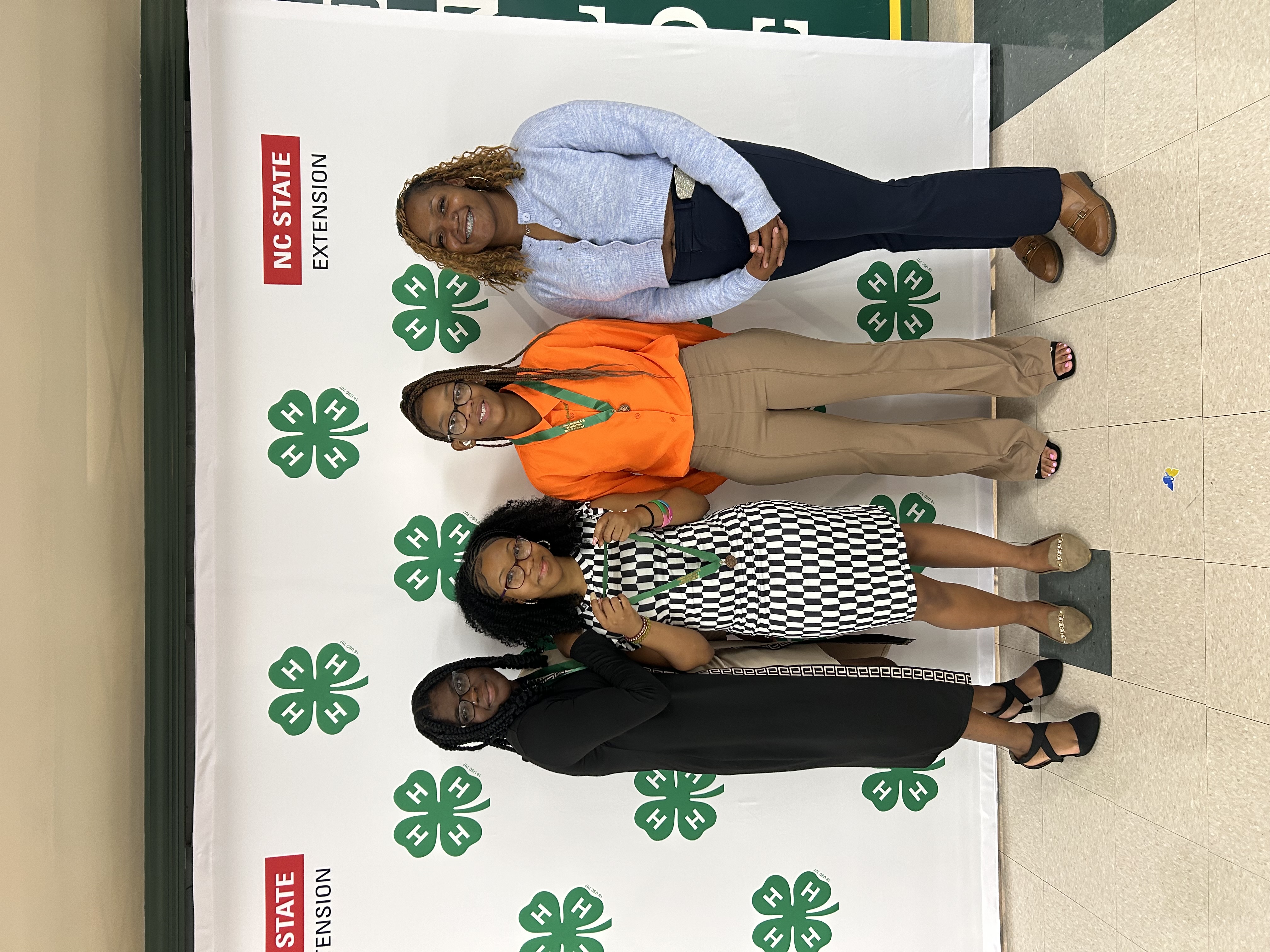 Four girls - two wearing dresses and two wearing dress pants - stand in front of a white background decorated with the official 4-H clover emblem and the Cooperative Extension banner. Three girls are wearing bronze medals. 