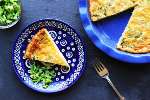 Top-down view of a golden-brown broccoli and cheese quiche in a blue ruffled pie dish, with one slice served on a matching decorative blue plate next to a silver fork and fresh broccoli.